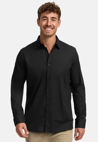 Hombre sonriente con cabello rizado, vistiendo una camisa negra de botones y pantalones beige, de pie contra un fondo claro y liso.