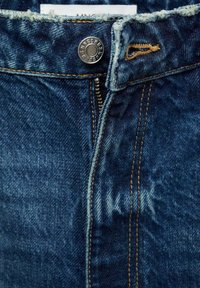 Un jean en denim bleu foncé avec une fermeture à bouton, une braguette zippée et des coutures contrastantes en tan le long des coutures. Détail de l'ourlet effiloché à la taille.