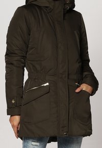 Parka isolée vert olive avec fermeture éclair sur le devant, taille réglable avec un cordon, grandes poches latérales et capuche. Présente des accents de boutons et de fermeture éclair.