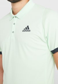 Polo vert clair en tissu texturé, avec une patte de boutonnage à trois boutons et des accents noirs sur les manches. Présente le logo Adidas sur la poitrine.