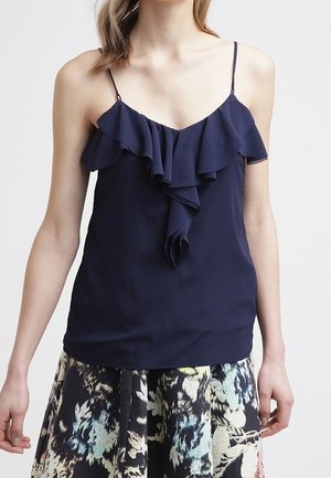 Frau trägt ein marineblaues ärmelloses Oberteil mit Rüschendetail vorne und dünnen Trägern, kombiniert mit einem hoch taillierten Floralen Rock.