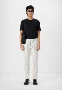 Schwarzes T-Shirt mit kurzen Ärmeln, hellgraue Cargo-Hose mit seitlichen Taschen, schwarze Umhängetasche und schwarze Sneaker, getragen von einer stehenden Figur.