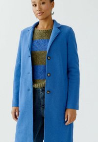 Blauwe wolmix jas met een enkele knoopsluiting, notch-kraag en een zachte textuur; gedragen over een olijf- en blauw gestreept sweater.