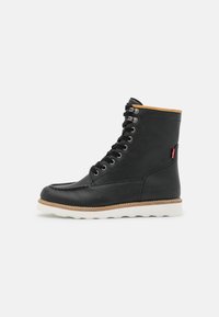 Botas de tobillo negras de cuero con un acabado texturizado, punta redonda, suela de goma blanca, acentos de cuero marrón y ojales de metal para los cordones.