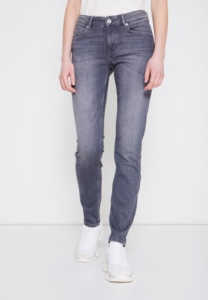 Persona con jeans skinny desgastados en gris y zapatillas blancas, de pie contra un fondo blanco liso.