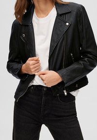 Schwarze Lederbikerjacke mit silbernen Reißverschlüssen und Druckknöpfen, taillierte Passform und Schulterklappen, kombiniert mit einem weißen T-Shirt und schwarzen Jeans.