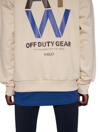 Beige sweatshirt met de tekst "AW OFF DUTY GEAR 223 AALBORG TACTICAL WING HALO", gedragen over een blauw overhemd, met ontspannen handen langs de zij.