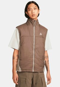 Brun isolerad väst med hög krage, dragkedja fram och grå detaljer. Har en logotyp på bröstet och bärs över en ljus skjorta.