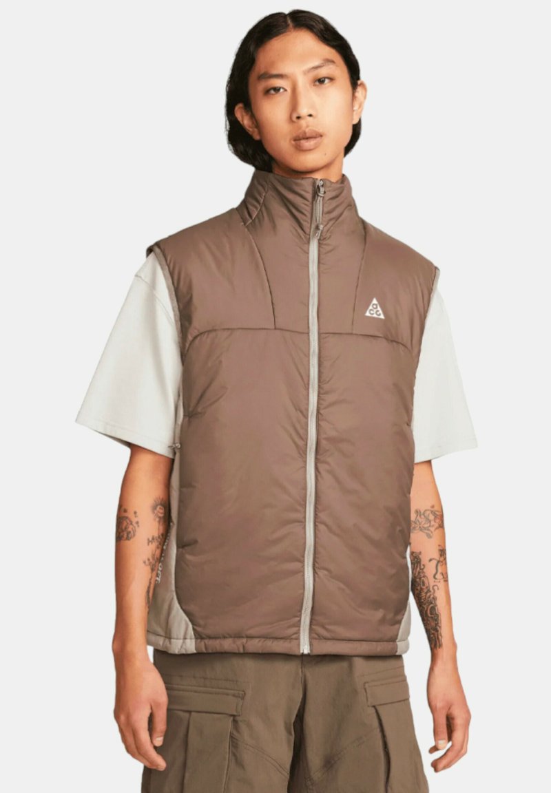 Brun isolerad väst med hög krage, dragkedja fram och grå detaljer. Har en logotyp på bröstet och bärs över en ljus skjorta.