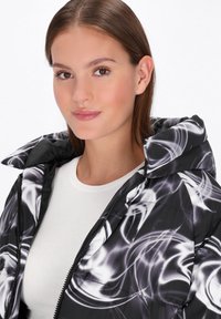 Sort pufferjakke med hvidt abstrakt swirls mønster, der har hætte, lynlås og en glat, blank overflade. Hvid bluse under.