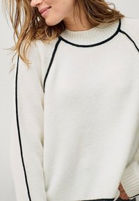 Maglione lavorato a maglia in crema con dettagli neri lungo il collo e le cuciture laterali, caratterizzato da un colletto a coste e una vestibilità comoda. Morbido al tatto.