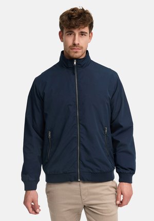 Mężczyzna ubrany w granatową bluzę na zamek z wysokim kołnierzem oraz beżowe spodnie na jasnym, jednolitym tle.