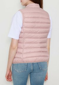 Gilet rembourré rose clair avec un design matelassé, col haut et sans manches, porté sur un t-shirt blanc et associé à un jean bleu.
