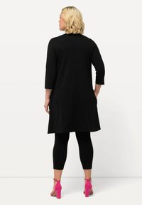 Schwarzes Tunika-Kleid mit drei Viertelärmeln, knielang, kombiniert mit schwarzen Leggings und pinken High-Heels. Glatte Textur.