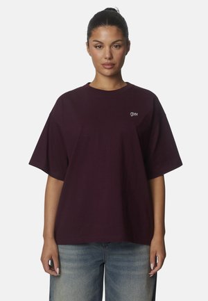 Donna con capelli scuri raccolti in uno chignon indossa una t-shirt larga viola scuro a maniche corte con una piccola scritta bianca sul petto e jeans blu sbiaditi.