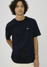 Jeune homme aux cheveux bouclés portant un T-shirt bleu marine uni avec un petit logo doré brodé sur la poitrine, debout devant un fond uni.