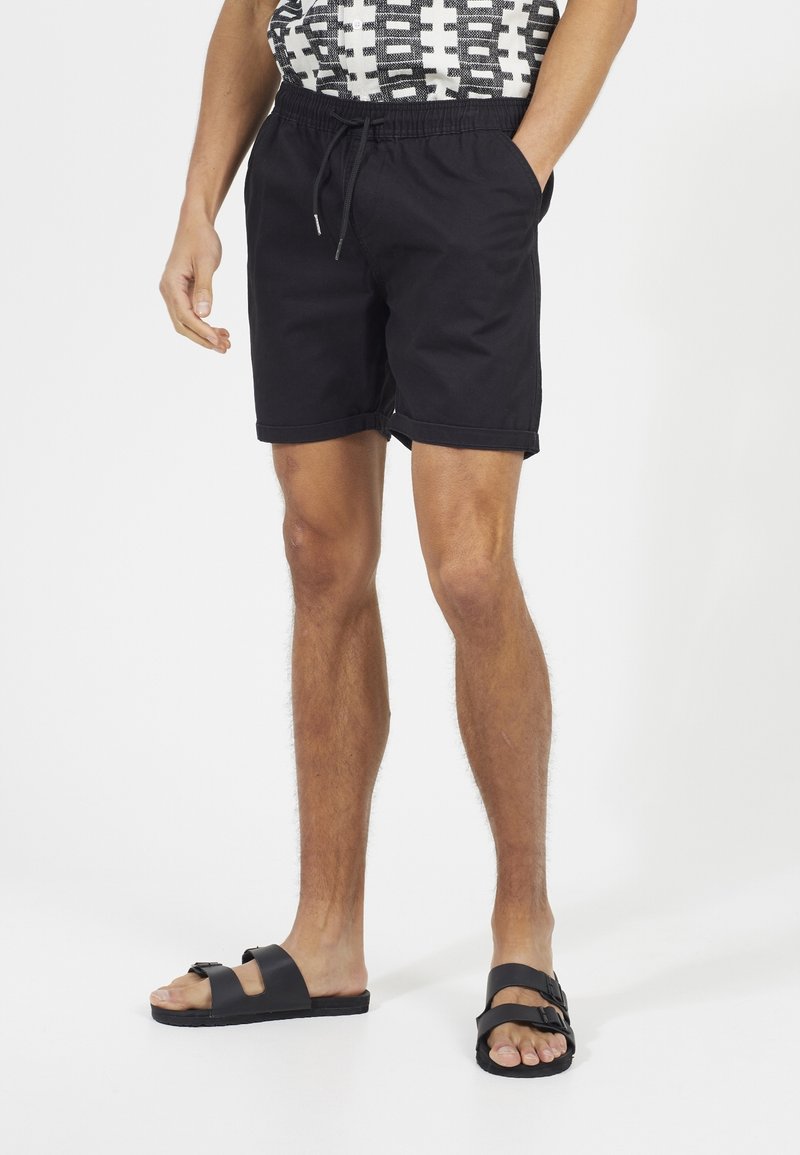 Czarne kąpielówki z elastycznym pasem i sznurkiem, charakteryzujące się prostą, gładką teksturą oraz luźnym, swobodnym krojem. Noszone z czarnymi sandałami.