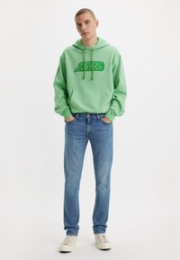 Νεαρός άνδρας φορώντας ανοιχτό πράσινο φούτερ με το λογότυπο "Sequoia", μπλε τζιν και λευκά αθλητικά παπούτσια, στέκεται με τα χέρια στις τσέπες σε λευκό φόντο.