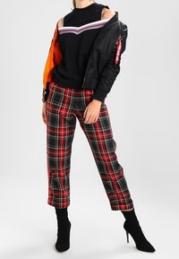 Kvinna iklädd svart cold-shoulder topp, svart bomberjacka delvis av, röd rutig byxor och svarta högklackade ankelstövlar.