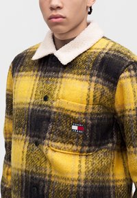 Млад човек носи жълто и черно карирано яке с бял пухен яка и лого Tommy Jeans на джоба на гърдите.