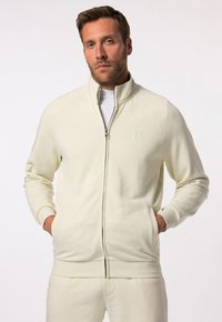 Sudadera de cremallera color beige claro con cuello alto, puños acanalados y logo bordado. Textura suave, diseño ajustado y bolsillos laterales.