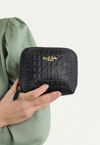 Portafoglio in pelle nera con incisione a coccodrillo, con branding dorato, chiusura a zip e superficie testurizzata. Tenuto in una mano che indossa una maglietta verde.