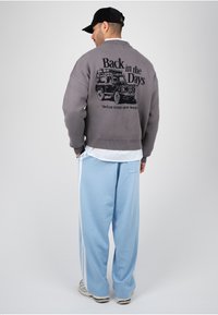 Homme portant un pull gris avec "Back in the Days" et un motif représentant un véhicule, pantalon bleu clair à rayures blanches, casquette noire et baskets, de dos.