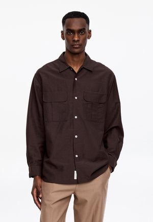 Mężczyzna ubrany w ciemnobrązową koszulę na guziki z kieszeniami na piersi oraz beżowe spodnie, stojący na tle jednolitego białego tła.