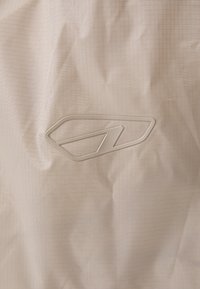 Beige stof med et rutemønster, der har et hævet, hvidt logo. Materialet ser glat ud med en let rynket struktur.