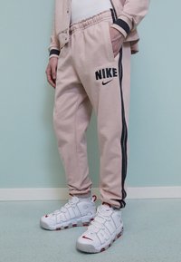 Ljusrosa träningsbyxor med svarta sidostreck och en stor svart "Nike"-logotyp. I kombination med vita sneakers med röda accenter och snörningsdesign.