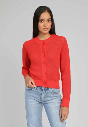Jeune femme portant un cardigan rouge vif boutonné et un jean bleu clair, se tenant devant un fond gris uni.
