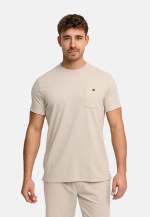 Homme portant une chemise beige à manches courtes texturée et un pantalon assorti, face à l'avant sur un fond blanc uni.