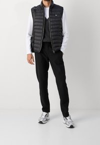Hombre que lleva un chaleco acolchado negro, un chaleco de punto gris, una camisa de manga larga blanca, pantalones negros, zapatillas blancas y lleva una mochila negra.
