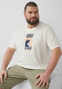 Lichtbeige t-shirt met korte mouwen, voorzien van een grafische print met blauwe en perzikkleurige accenten. G gedragen met olijfgroene shorts, met een relaxte pasvorm.