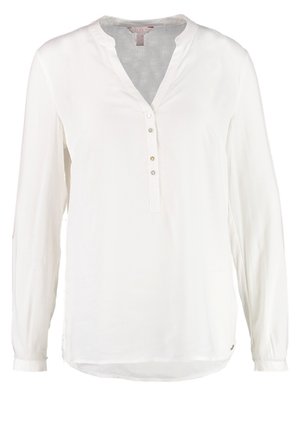 Frau trägt eine weiße Bluse mit langen Ärmeln, schwarze Hose und schwarze genoppte Oxford-Schuhe, geht vor einem schlichten hellen Hintergrund.