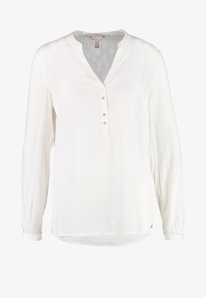 Vrouw draagt een witte blouse met lange mouwen, zwarte broek en zwarte gestekelde oxford schoenen, lopend tegen een effen lichte achtergrond.