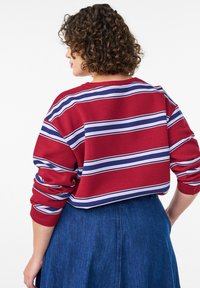 Červený pletený sveter s horizontálnymi bielymi a modrými pruhmi, s voľným strihom a rebrovanými manžetami, v kombinácii s modrou rifľovou sukňou.