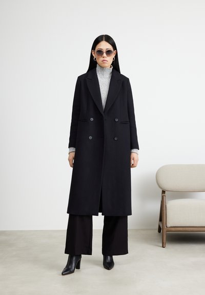 Cappotto nero doppiopetto, design sartoriale, con rever a punta e tasche laterali. Il modello indossa un dolcevita grigio e pantaloni neri a gamba larga.