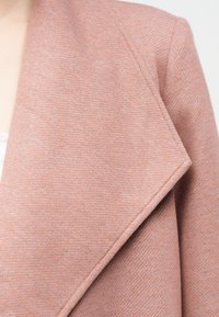 Gros plan sur une personne portant un manteau en laine rose poudré texturé avec un large revers cranté, par-dessus un haut de couleur claire.