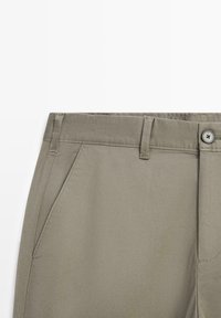 Gros plan sur la ceinture d'un pantalon beige avec passants pour ceinture, poche latérale, fermeture par bouton et braguette éclair sur fond blanc.