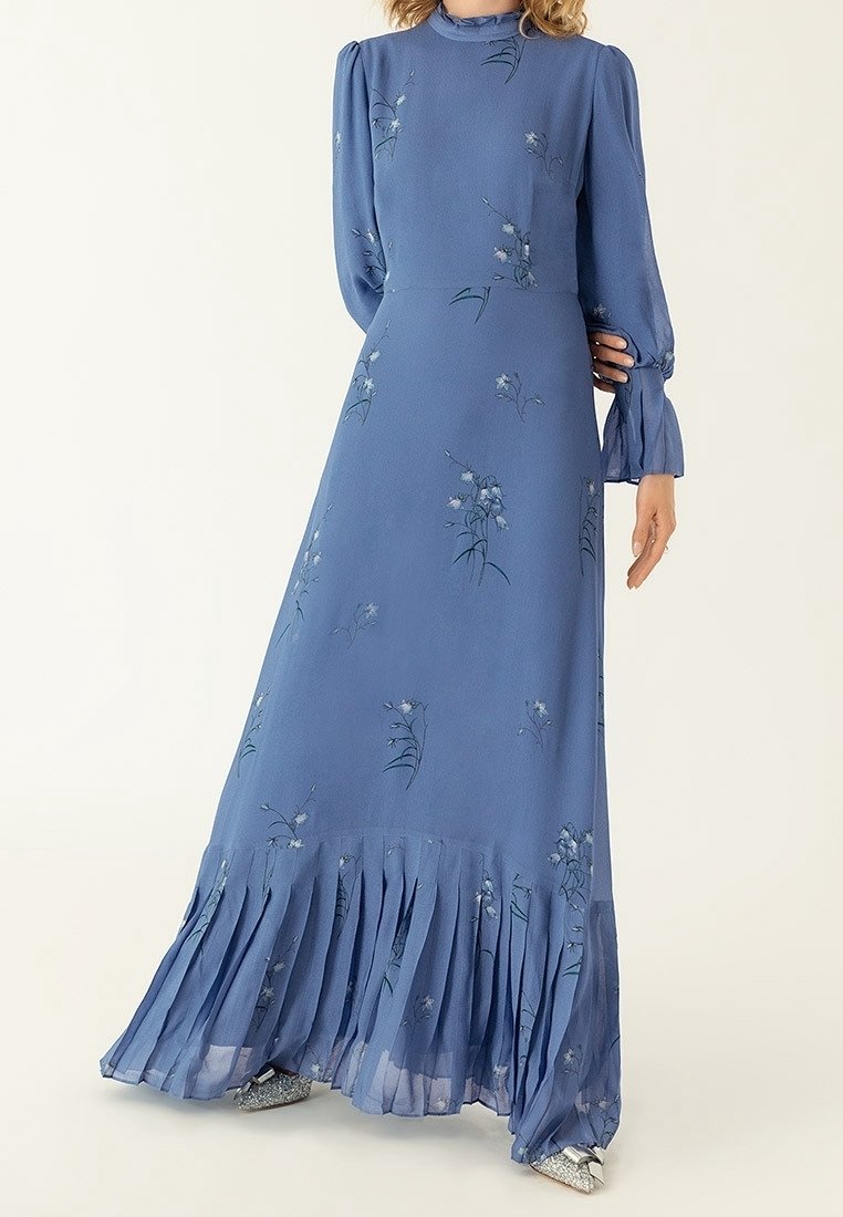 Femme portant une robe bleue à manches longues, col montant, imprimé floral et ourlet plissé, debout avec une main sur la hanche.
