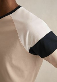 Īsām piedurknēm t-krekls ar gaiši bežīgu korpusu un tumši z blue piedurknēm. Gluda auduma, ar apaļu kakla izgriezumu un kontrastējošiem plecu šuvēm.