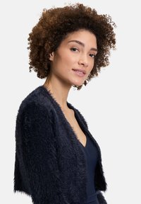 Veste en fausse fourrure marine avec un design court, devant ouvert et surface texturée. Caractéristiques d'un matériau doux et duveteux avec un col simple.