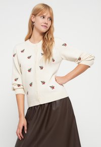 Κρεμ ζακέτα με κεντήματα λουλουδιών με κόκκινους μούρους και πράσινα φύλλα, κλείσιμο με κουμπιά και πλεκτές μανσέτες και τελείωμα. Ορατή καφέ φούστα.