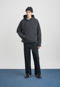 Nuori henkilö, jolla on musta pipo, tummanharmaa huppari, mustat housut ja mustat lenkkarit, seisoo sinisellä matolla valkoista seinää vasten.