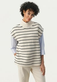 Pulover fără mâneci din tricot, de culoare crem, cu dungi orizontale navy, gât înalt și o croială lejeră. Asortat cu o cămașă albastru deschis și pantaloni bej.