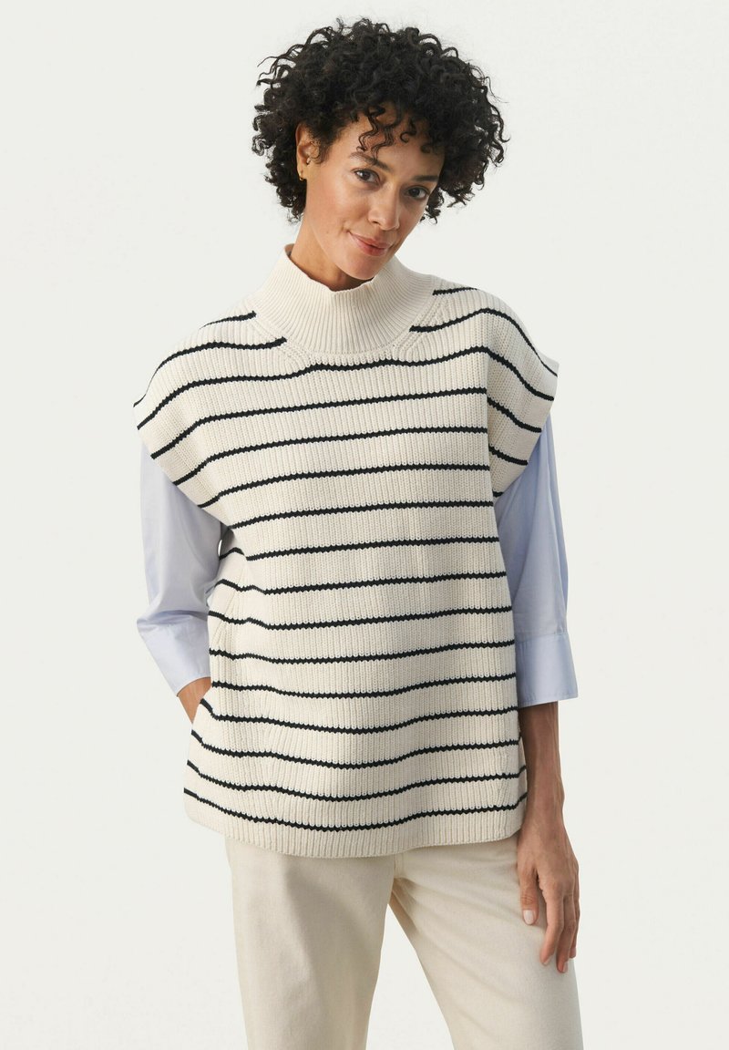 Gehaakte mouwloze trui in roomkleur met marineblauwe horizontale strepen, hoge hals en losse pasvorm. Gepaard met een lichtblauw shirt en beige broek.