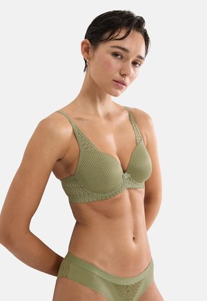 Vrouw met kort donker haar, gekleed in een olijfgroen kanten beha en bijpassende slip, staande tegen een effen lichte achtergrond met een neutrale gezichtsuitdrukking.