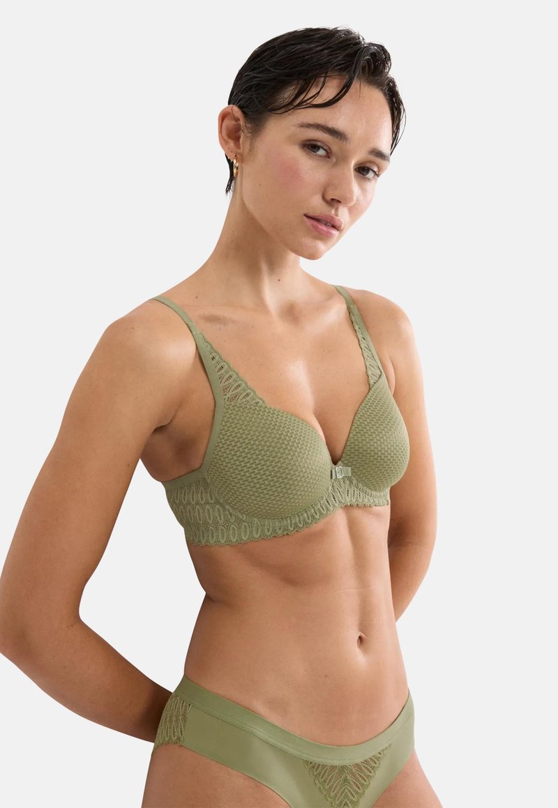 Γυναίκα με κοντά σκούρα μαλλιά που φοράει εσώρουχα δαντέλας σε ελιά πράσινο, στέκεται μπροστά σε απλό φωτεινό φόντο με ουδέτερη έκφραση.