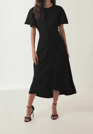 Femme portant une robe midi noire à manches courtes et taille froncée, associée à des sandales à talons hauts noires avec bride à la cheville, sur un fond uni.